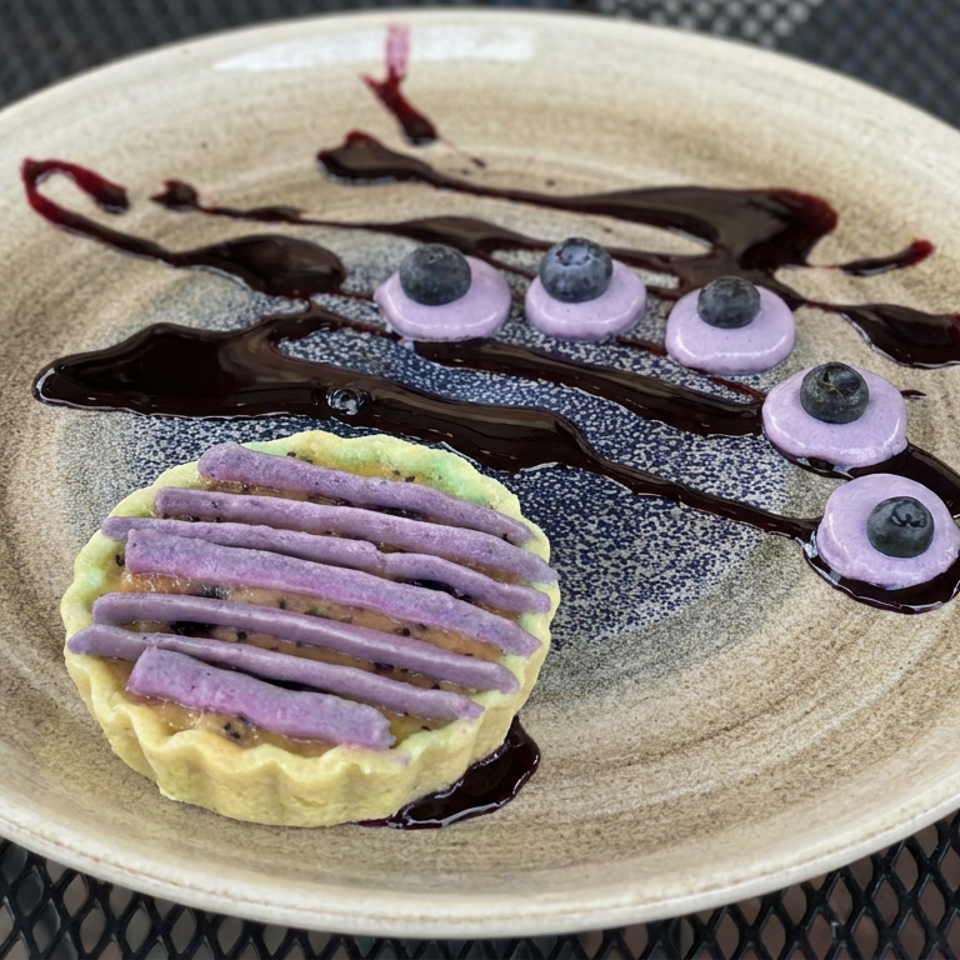 Decorative dessert plate with a tart topped with purple cream, blueberries, and chocolate sauce.